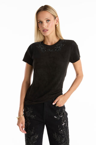 A woman with straight blonde hair wears the Amaya Floral Sequin T-Shirt in black, featuring beaded embellishments near the neckline, paired with matching black pants with floral embroidery, standing against a plain white background.