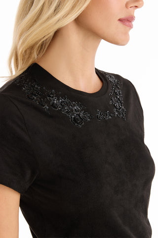 A woman with loose blonde hair is shown in profile, wearing the Amaya Floral Sequin T-Shirt in black, which features intricate floral sequin embroidery along the neckline. Only her face and upper torso are visible.