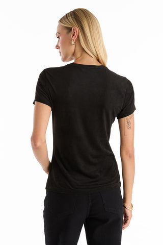 A woman with straight blonde hair, hoop earrings, and a small tattoo on her left upper arm stands facing away from the camera, wearing black pants and the Amaya Floral Sequin T-Shirt in black.