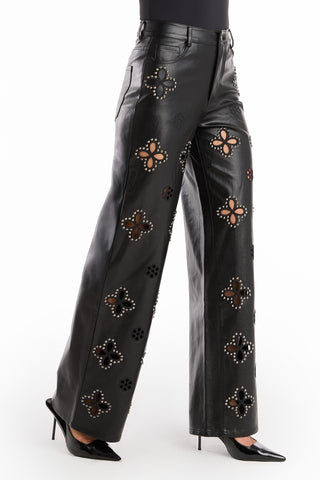 Someone wears the Kaia Studded High Rise Leather Pant in black, featuring large floral cutouts and silver studs, paired with black pointed high heels against a white background.