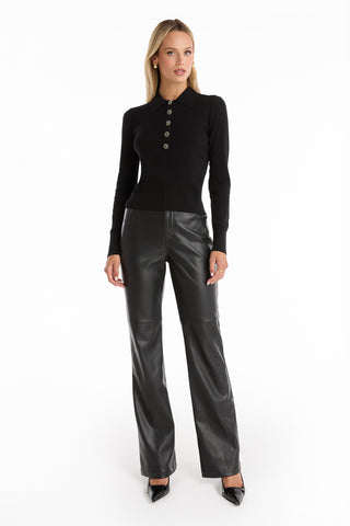 A woman with long blonde hair and a neutral expression models the Darla Rose Polo Shirt - Black against a white background, paired with high-waisted black leather pants and black pointed-toe heels for a look of classic sophistication.