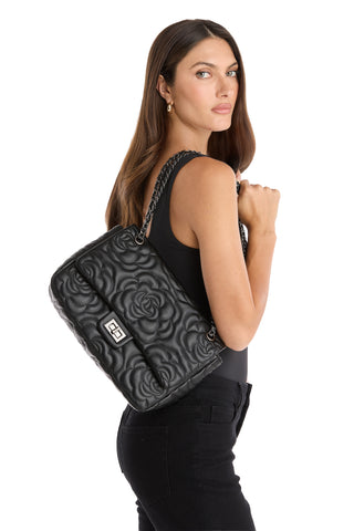 A woman with long brown hair, dressed in a black sleeveless top and black jeans, carries the Alessia Rose Embossed Leather Bag - Black with a chain strap over her shoulder, standing against a white background.
