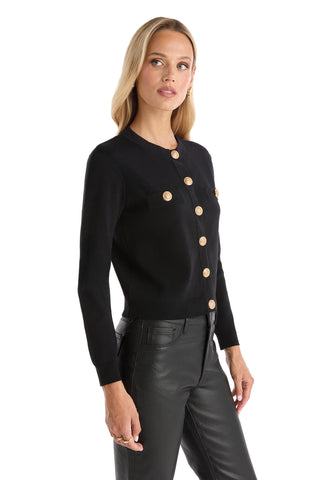 A woman with long blonde hair wears the Loretta Knit Cardigan Top in black, featuring gold dome buttons and chest pockets, paired with black leather pants. She stands against a white background, looking slightly to the side.