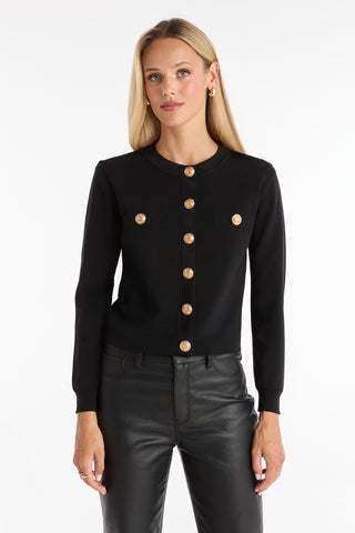A woman with long blonde hair wears the Loretta Knit Cardigan Top in black, featuring padded shoulders, gold buttons, and front pockets, paired with black leather pants against a plain white background.