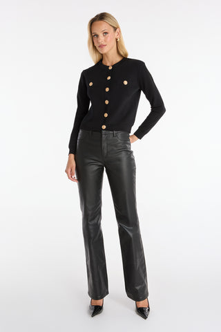 A woman with blonde hair stands against a white background wearing the Loretta Knit Cardigan Top in black, featuring large gold buttons and padded shoulders, paired with black leather pants and pointed-toe heels.