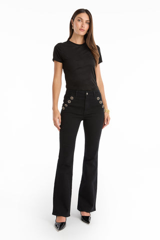 A woman stands against a white background wearing a fitted black t-shirt, Allegra Rose Jean - Black high-waisted flared jeans with silver grommets on the hips, black pointed-toe heels, and gold bracelets.