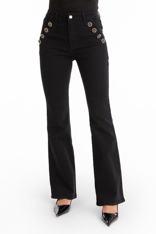 A person models the Allegra Rose Jean - Black, featuring high-waisted flare legs and decorative rose buttons, paired with black pointed-toe shoes against a white background.