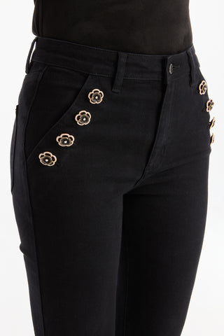 Close-up of a person wearing Allegra Rose Jean - Black, featuring metallic floral embellishments on the pockets and paired with a black top, against a plain white background.