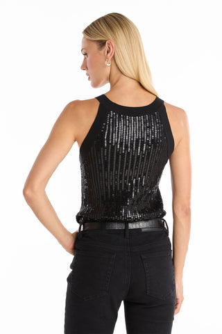 A woman with straight blonde hair stands facing away with one hand on her hip, wearing the Shae Sequin Halter Tank - Black and black jeans, accessorized with a black belt and silver hoop earrings for a chic, modern look.