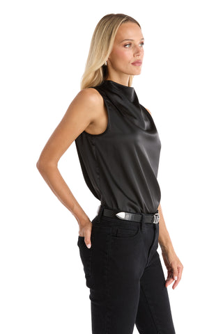 A woman with long blonde hair wears the Mara Silky Top - Black, a sleeveless mock neck satin blouse, tucked into black jeans with a black belt, standing against a white background.