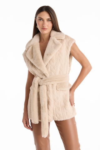 A woman stands confidently against a white background, wearing brown leather pants, a beige faux fur belted vest, and the Leone Cable Knit Cardigan in Pearl draped casually over her shoulders.
