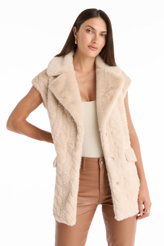A woman with straight brown hair wears a sleeveless beige faux fur vest over a light top and tan leather pants, standing with one hand on her hip; the Leone Cable Knit Cardigan - Pearl is draped casually over her arm.