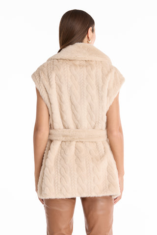 A woman with straight brown hair is seen from the back wearing the Leone Cable Knit Cardigan in Pearl, a beige textured knit with a tie-front waist, paired with tan leather pants. The backdrop is white.