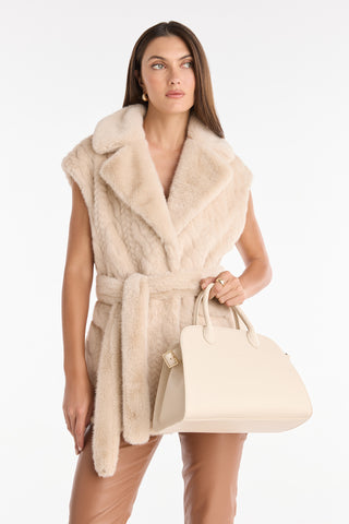 A woman in a sleeveless beige faux fur vest with a belt, tan leather pants, and holding the Margo Leather Bag - Pearl poses against a plain white background.