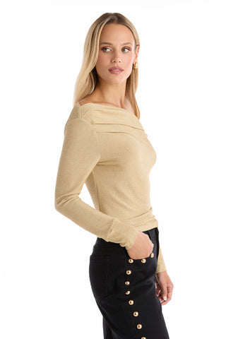 A woman with long blonde hair wears the Victoria Shimmer Top - Gold, a fitted long-sleeve top, paired with black pants featuring gold side buttons. She is shown in profile against a white background, glancing over her shoulder.