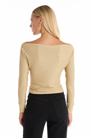 A woman with straight blonde hair, seen from behind, wears the Victoria Shimmer Top in gold with off-the-shoulder long sleeves and black jeans against a white background.