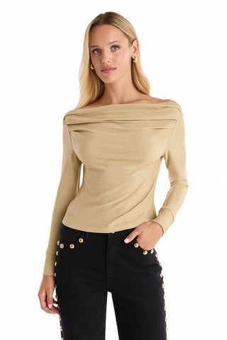 A woman with long blonde hair wears the Victoria Shimmer Top - Gold with long sleeves and a boat neck, paired with black pants featuring gold buttons, standing against a white background.
