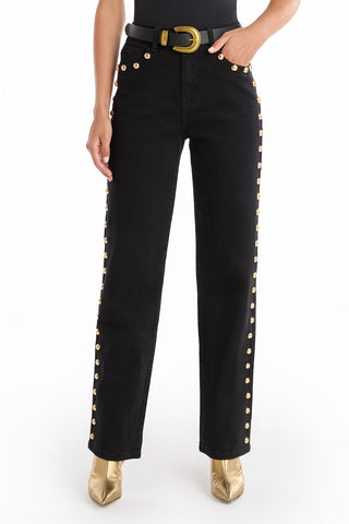 Person wearing Leah Studded Jean - Black, a high-rise four-way stretch denim jean with gold buttons along the side seams, paired with a black belt featuring a large gold buckle and gold pointed-toe boots. Upper torso and head not shown.