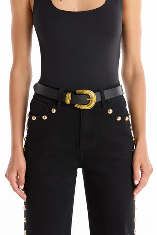 A person wears a sleeveless black top and high-waisted black jeans with gold studs, accessorized with the Shelly Belt - Black, which features a large gold cowboy buckle for a western-inspired look.
