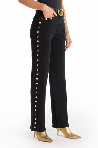 The lower half of a person is shown wearing the Leah Studded Jean - Black, a high-rise denim with gold button accents on the pockets and sides, styled with gold pointed-toe heeled boots and a black belt.
