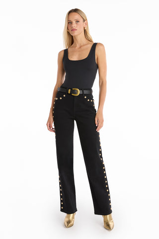 A woman stands against a white background wearing a black sleeveless top, Leah Studded Jean - Black with gold grommet details, a black belt with a gold buckle, and gold ankle boots.