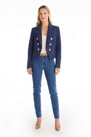 A woman stands facing forward against a white background, wearing the Gabrielle Double Breasted Blazer - Navy with gold buttons, a white top, blue jeans, a beige belt, and beige heels.