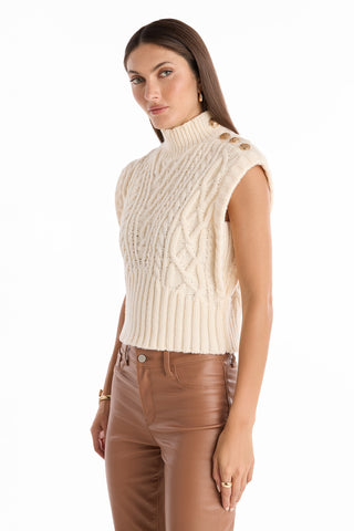 A woman wears the Raina Cable Knit Button Sweater in Pearl—sleeveless with a mock turtleneck and shoulder buttons—paired with high-waisted brown leather pants, standing against a white background.