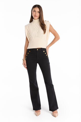 A woman stands against a white background wearing a sleeveless cream cable-knit turtleneck sweater, Allegra Bootcut Jean in black with gold buttons, and beige pointed-toe heels.
