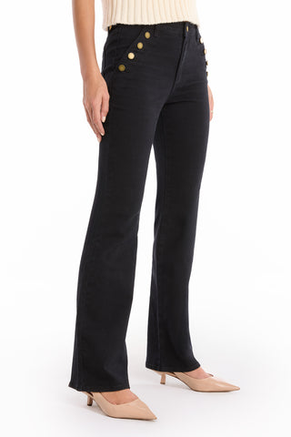 Wearing the Allegra Bootcut Jean in black, styled with gold-button front pockets, a cream sleeveless top, and beige low-heeled shoes, a person stands against a white background for a vintage-inspired look.