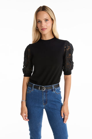 A woman with long blonde hair wears the Sylvie Lace Sleeve Top - Black, with lace puff sleeves and a knit crewneck, paired with blue jeans and a black belt, standing against a white background.