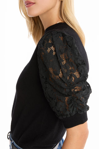 A woman wearing the Sylvie Lace Sleeve Top - Black, featuring sheer lace puff sleeves and a knit crewneck, paired with blue jeans. She is shown from the side with her face partially visible.