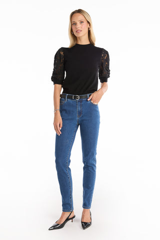 A woman stands against a white background wearing the Layla Skinny Jean in medium blue, a black lace puff sleeve top, black belt, and black pointed-toe heels. She has straight blonde hair and a relaxed expression.