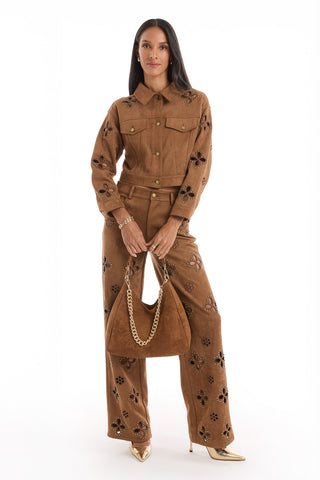 A woman faces forward in a brown suede two-piece with floral cut-outs and gold buttons, holding the Tribeca Chain Suede Bag - Cognac with a gold chain handle. She pairs it with matching pants and gold pointed-toe heels.