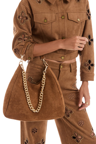 A person in a brown suede jacket and floral cutout pants holds the Tribeca Chain Suede Bag - Cognac, which features a gold chain strap.