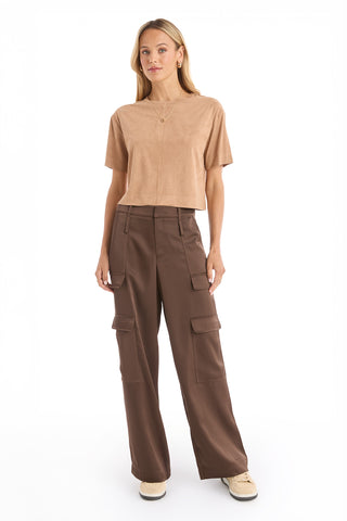 A woman stands against a white background wearing the Naya Suede T-Shirt - Cafe with a cropped fit, paired with dark brown wide-leg cargo pants, beige sneakers, and simple jewelry.