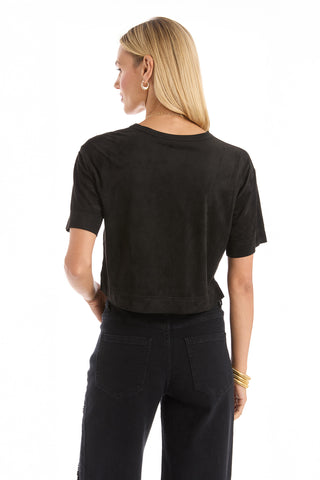 A woman with long blonde hair models the Naya Suede T-Shirt - Black in a cropped fit and black jeans, standing with her back to the camera against a white background. She styles the look with gold hoop earrings and bangles.