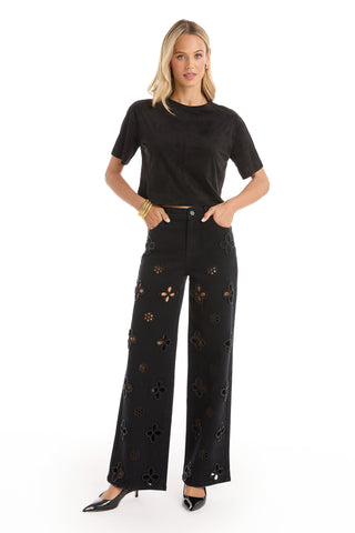 A woman stands against a white background, wearing the Naya Suede T-Shirt in black as a chic layering piece, paired with black wide-leg pants featuring cutout floral patterns, black heels, and gold bracelets, hands in her pockets.