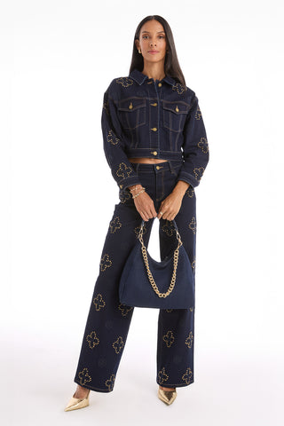 A woman faces forward in a matching dark denim jacket and pants with gold studs, gold heels, and holds the Tribeca Chain Denim Bag - Indigo featuring a gold chain strap. The background is white.