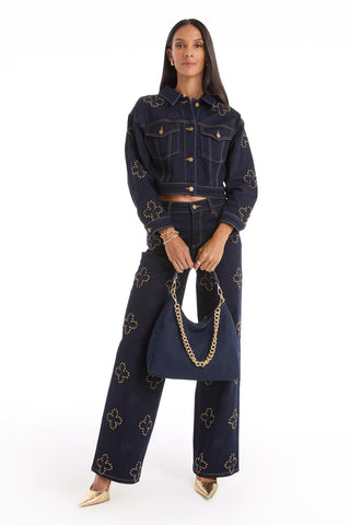 A woman faces forward in a dark denim jacket and wide-leg pants with gold embellishments, holding the Tribeca Chain Denim Bag - Indigo with bold gold chain detail, and wearing gold heels against a white background.