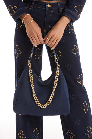 A person in dark blue studded denim pants and jacket holds the Tribeca Chain Denim Bag - Indigo, featuring a thick gold chain strap, with gold rings and bracelets. The background is white.