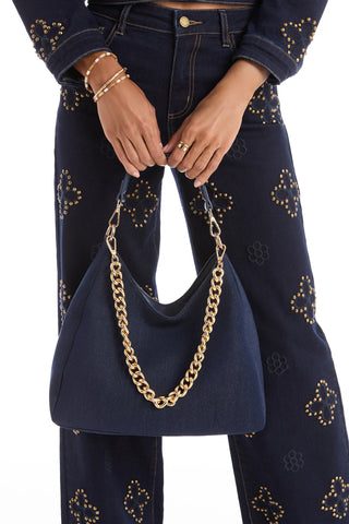 A person in dark blue studded denim pants and jacket holds the Tribeca Chain Denim Bag - Indigo, which has a thick gold chain detail. Their hands and wrists are accessorized with gold rings and bracelets.