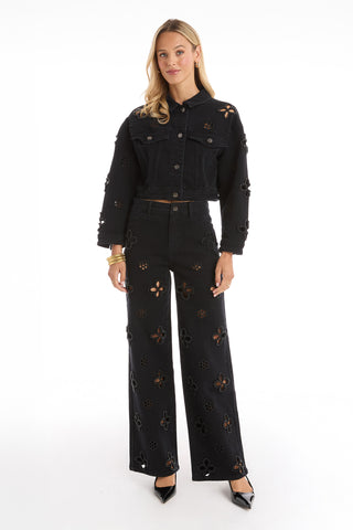 A woman with long blonde hair faces forward in the Kaia Pearl Jean Jacket - Black and wide-leg pants, both with floral cutout designs. She wears black heels and gold bangles, set against a plain white background.