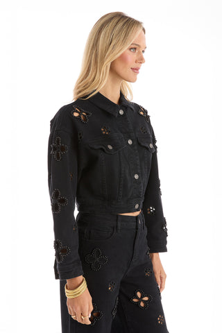 A woman with long blonde hair wears the Kaia Pearl Jean Jacket in black, paired with matching high-waisted black jeans featuring floral cut-out designs. She stands in profile against a white background and accessorizes with gold bangles.