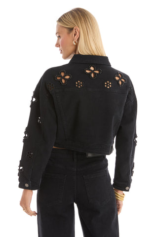 A woman with shoulder-length blonde hair and gold jewelry faces away, wearing the Kaia Pearl Jean Jacket in black—an embellished cropped denim piece with floral cut-outs—paired with matching high-waisted jeans.