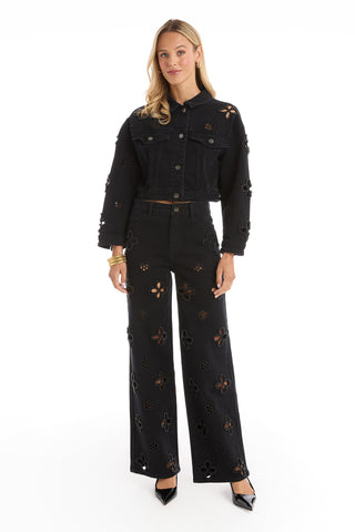 A woman poses confidently in the Kaia Pearl Jean Jacket - Black, paired with matching high-waisted pants. She has long blonde hair, wears black heels and gold bracelets, all set against a white background.
