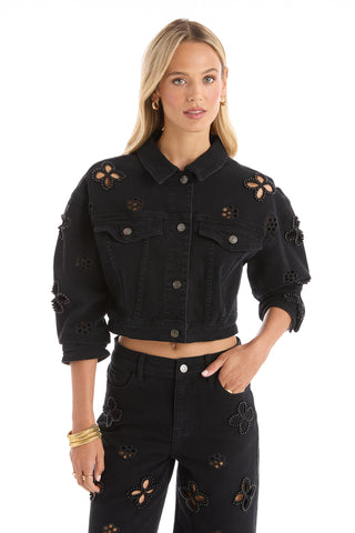 A woman with long blonde hair wears the Kaia Pearl Jean Jacket in black with gold hoop earrings and bangles, posing stylishly against a white background.