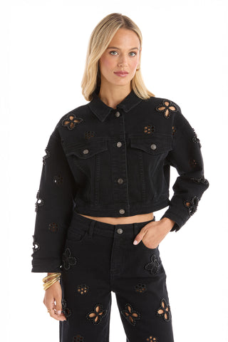 A woman wears the Kaia Pearl Jean Jacket in black and matching cut-out floral jeans, posing with one hand in her pocket against a plain white background.