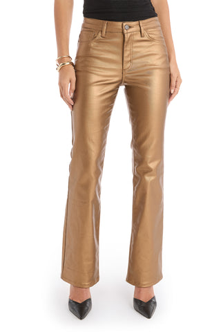 A woman stands against a white background wearing Aria Coated Flare Jean - Gold Metallic with a high-rise fit, paired with a black top, black pointed shoes, and gold bracelets.
