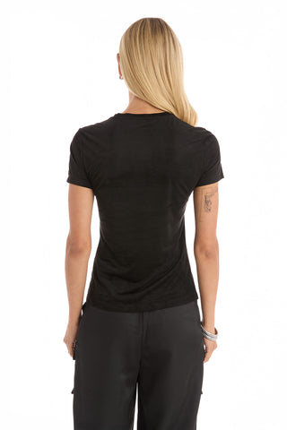 A person with long blonde hair, wearing the Amaya Suede T-Shirt in Black and dark pants, stands facing away from the camera against a white background.