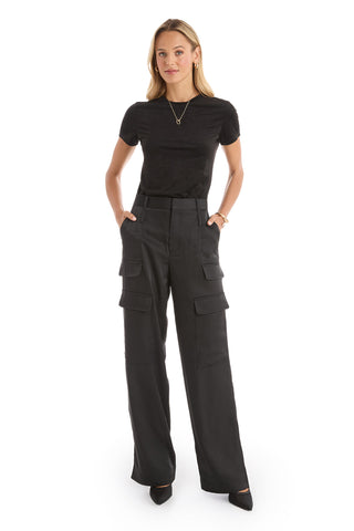 A woman faces forward wearing the Amaya Suede T-Shirt in black, paired with high-waisted, wide-leg black cargo pants with side pockets. Her hands are in her pockets and she completes the look with black pointed-toe shoes.